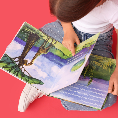 A little girl reading a picture book on her lap. The book has a blue sky, giraffe and trees. 