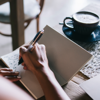 Someone writing in a grid notebook with a cup of coffee set aside.
